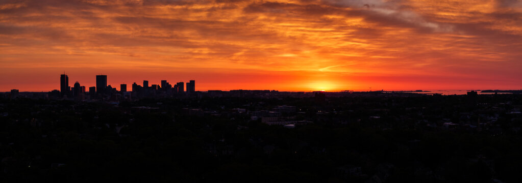 Aerial view of silhouetted skyscrapers punctuate the horizon beneath a fiery dawn sky, the city awakening beneath a blanket of warm, orange hues, Boston, Massachusetts, United States.