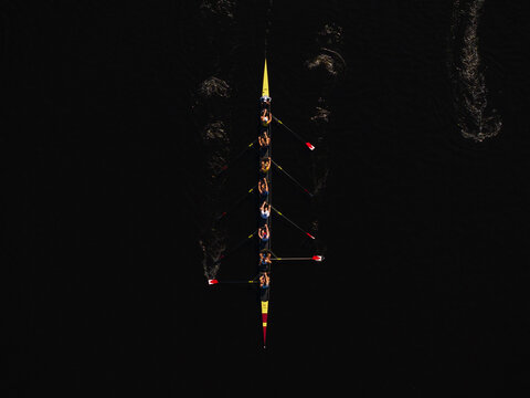 Aerial view of a rowing team slicing through the dark waters of the Charles River, their oars flashing red and yellow against the inky blackness, Boston, Massachusetts, United States.