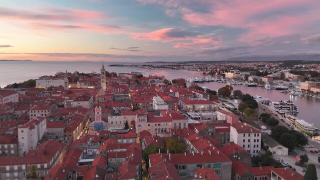 Drone flies over Old Town Zadar in soft evening light, revealing pink sky, red rooftops, St. Donatus church and the calm Adriatic Sea. Autumn.