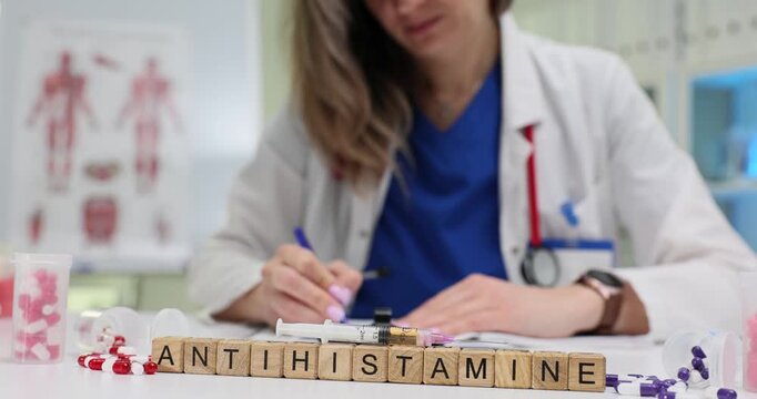 Doctor sits at desk writing detailed treatment plan for patient. Miniature wooden cubes create word Antihistamine on table near scattered tablets