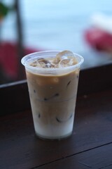 Refreshing iced coffee with milk in a clear plastic cup, sitting on a wooden table with ocean view in the background.