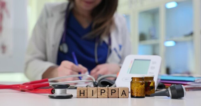 Wooden blocks spell word HIPPA near blood pressure monitor and brown vial. Woman doctor writes patient record guarding medical privacy rights