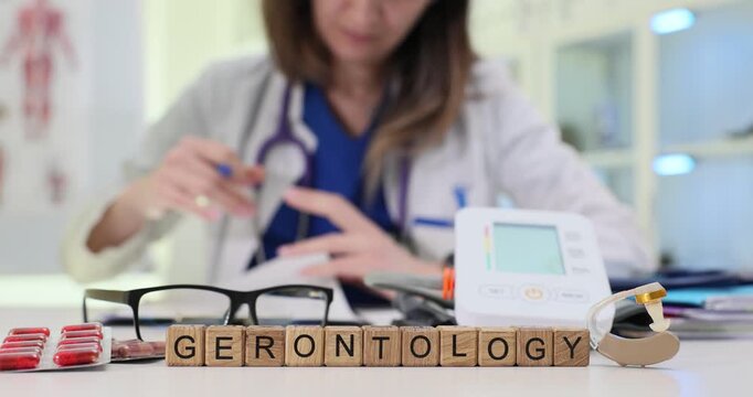 Wooden blocks spell word Gerontology near eyeglasses and blood pressure monitor. Woman doctor reviews aging research notes designing care plan