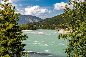 Turquoise river winds through the valley in Vinstra Innlandet Norway with mountains in the background. Green trees frame the scene as clouds float above