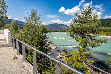 Scenic viewpoint by Gudbrandsdalsl&aring;gen river in Vinstra, Norway. Lush green trees line the riverbank, while mountains rise in the distance under a clear blue sky