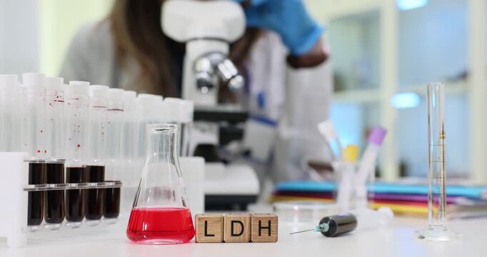 Wooden blocks spell short term LDH near red reagent flask and blood tubes. Laboratory doctor studies enzyme level recording test result in notebook