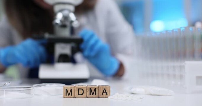 Wooden blocks spell word MDMA near white pills and Petri dishes. Scientist in blue gloves checks sample under microscope searching dangerous traces
