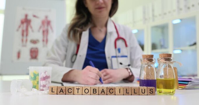Wooden blocks spell word Lactobacillus near glass bottles with colored liquid. Woman doctor writes notes about probiotic bacteria and gut balance
