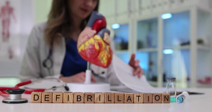 Wooden blocks spell word Defibrillation near heart model on table. Woman cardiologist reads ECG strip planning urgent electric shock treatment