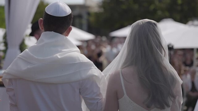 Rear view of a couple standing together under the chuppah during a traditional jewish ceremony. The bbride wearing a veil and the groom with a kippah take part in a cultural ritual.