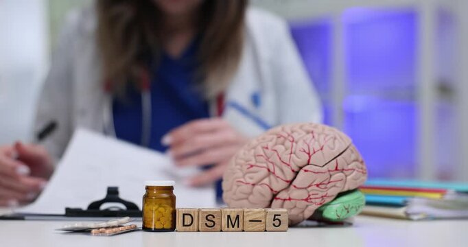 Wooden blocks spell code DSM 5 beside brain model on clinic table. Woman psychiatrist checks notes writing mental disorder diagnosis criteria