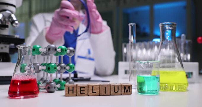 Wooden blocks on table spell word Helium beside colorful reagents. Chemistry scientist holds goggles near test tube and flasks in lab slow motion