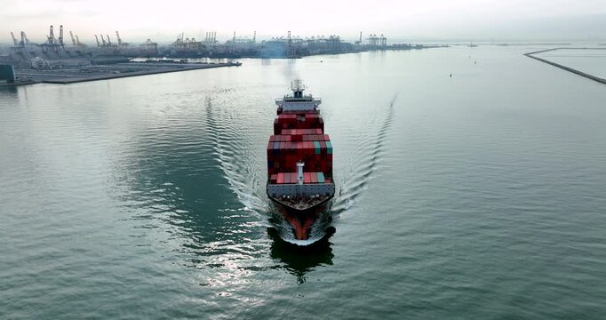 Aerial drone footage of a massive container ship fully loaded with cargo sailing across the ocean towards a busy industrial harbor, symbolizing global trade, logistics, and supply chain.