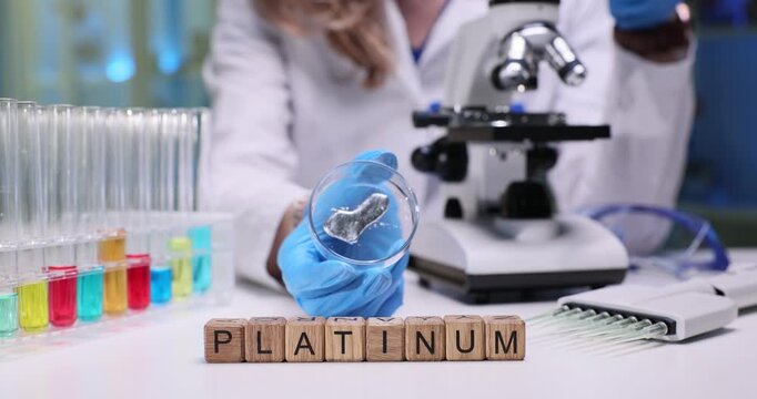 Wooden blocks spell word Platinum on desk with glass test tubes. Scientist in coat holds glass dish with metal sample near microscope on table