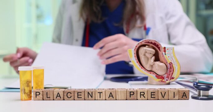 Wooden blocks on desk spell words Placenta Previa beside uterus model. Woman doctor writes patient pregnancy history while studying risk chart