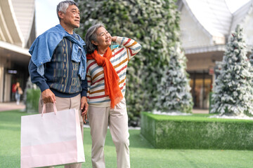 Happy senior asian couple holding shopping bag in front of snowy Christmas tree, enjoying winter holiday season, new year end sale festive shopping, senior lifestyle, love, joy and holiday celebration