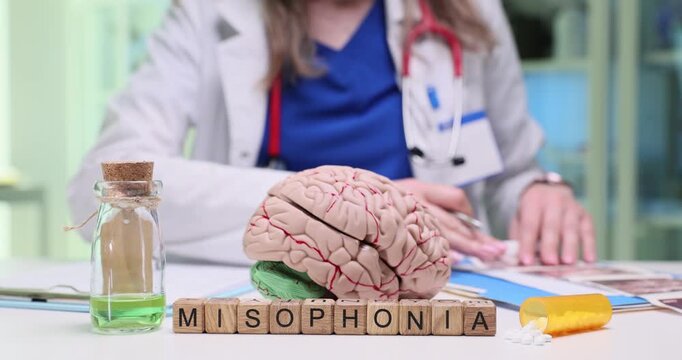 Wooden blocks spell word Misophonia near brain model on doctor desk. Woman neurologist writes notes about sound trigger disorder testing patient
