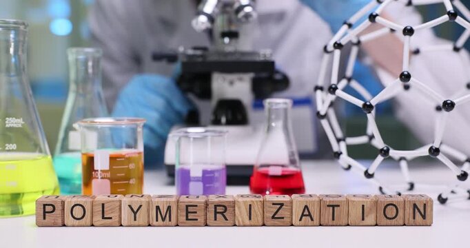 Wooden blocks on desk spell word Polymerization by bright flasks. Laboratory specialist monitors chain reaction progress noting results of microscope