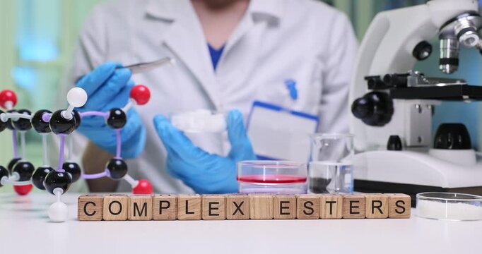 Laboratory technician conducts careful analysis of complex esters. Series of wooden blocks arranged near microscope create phrase Complex Esters