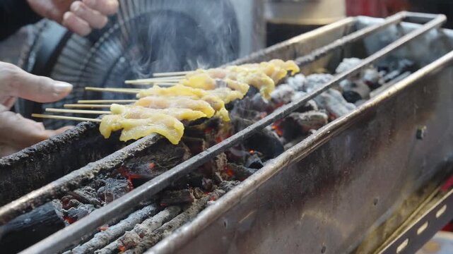 Sizzling pork or chicken satay being grilled on a traditional charcoal grill with smoke rising, a popular Thai street snack.