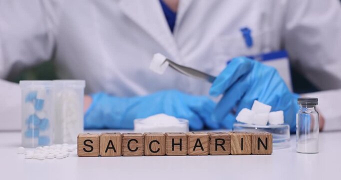 Lab technician holds small white sugar cube with pair of metal tweezers. Wooden blocks arranged neatly on table create word Saccharin as substitute