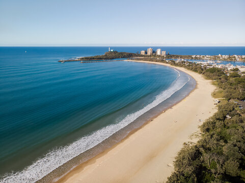 Aerial view of the sun-kissed beach curves gently toward the iconic Point Cartwright Lighthouse, where the azure ocean meets the golden sands, Mooloolaba, Queensland, Australia.