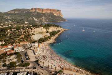 Aerial view of the vibrant beach packed with sunbathers under the gaze of the towering Cap Canaille cliff, its golden hues contrasting with the azure sea, Cassis, Provence-Alpes-Cote d Azur, France.
