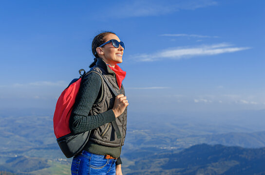 A happy young woman hiker wearing sunglasses and a backpack stands on a mountain peak against a clear blue sky, enjoying the scenic view.