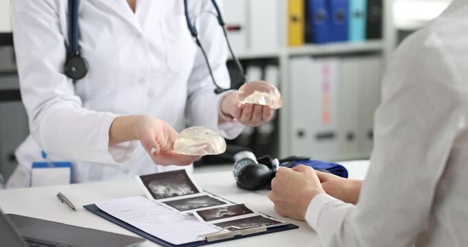 Woman doctor in white coat holds silicone breast implants during consultation. Doctor shows implants to lady patient explaining augmentation result