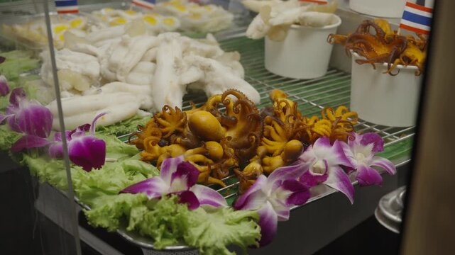 Close-up of fresh seafood skewers including grilled squid, garnished with flowers and small Thailand flags at SookSiam indoor market.