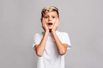 Surprised boy with hands on face looking at camera, emotional shocked child expressing amazement and astonishment on gray background, studio portrait of kid reacting with wide eyes and open mouth