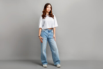 Beautiful full-length portrait of confident teenage girl with long dark hair standing in studio on gray background, looking at camera while wearing casual t-shirt and jeans
