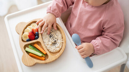 Child-friendly breakfast bear shaped plate with oats or porridge banana slices and assorted fresh fruits, vegetables and berries, served on a highchair tray. A kid eats using hand and silicone spoon