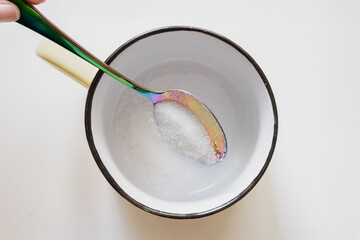 Putting tea spoon full of granulated sugar into cup, closeup top view photo. Rainbow spoon with sugar over enamel mug on white background, minimal kitchen scene