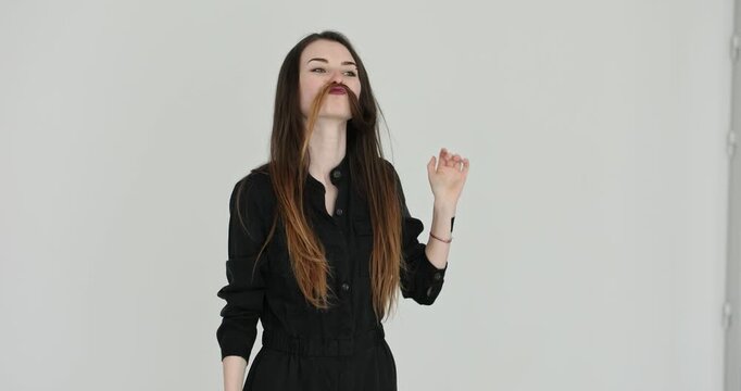 Young woman with long hair lifts strand as playful mustache. Woman poses in studio making funny expression during lighthearted joke slow motion