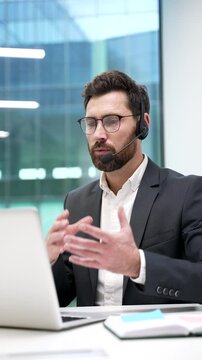 Businessman in a headset talking on a video call conference using laptop while sitting in office. The manager has a business meeting. Coach has online training, class, course or webinar. Vertical