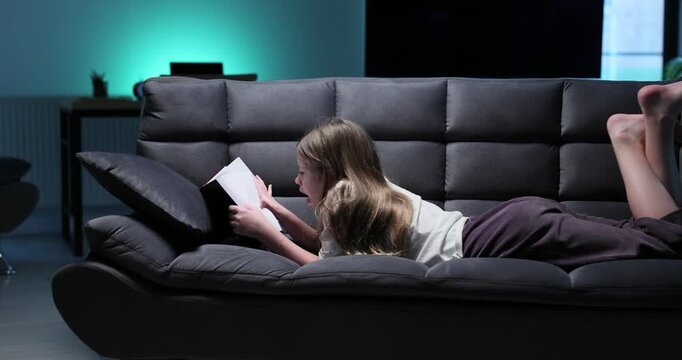 Young girl lies on dark sofa reading thick book under light. Teen girl kicks legs in air while staying absorbed in exciting adventure story pages