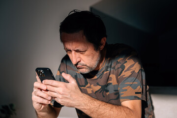 Mature man intensely focused on his smartphone, reading in concentrated silence as sunlight highlights his serious facial expression.