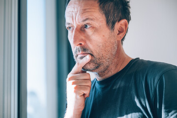 Authentic middle-aged caucasian man gazing thoughtfully out bright window, hand to mouth in deep concern. Soft natural daylight.