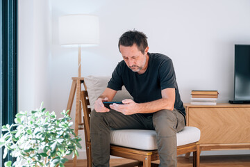 Caucasian man with beard sitting in modern living room, deeply concentrated on smartphone with...