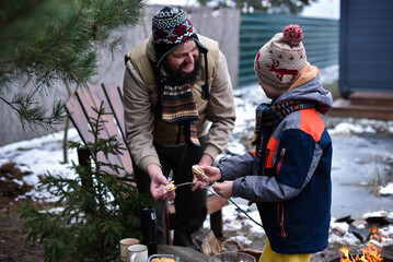 Smiling father and son in winter clothes holding roasted marshmallow on stick with cookies in backyard