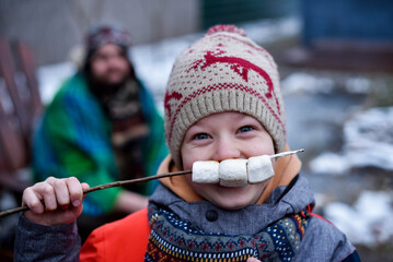 Funny boy in winter hat holding marshmallow on stick near face
