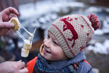 Joyfully surprised boy in winter clothes looking at roasted marshmallow in adult hands