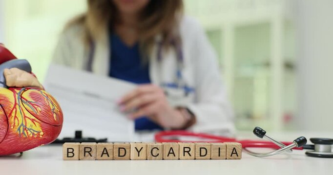 Wooden blocks spell word Bradycardia near heart model at desk. Woman cardiologist reads report while evaluating slow pulse problem of patient