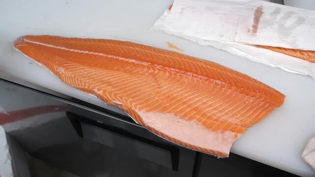 A professional chef prepares a raw salmon fillet on a white cutting board by meticulously using specialized fish tweezers to extract small pin bones from the orange flesh in a kitchen.