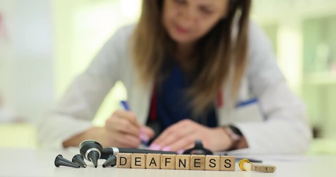 Wooden blocks on desk spell word Deafness near otoscope device. Woman doctor writes hearing test notes while reviewing audiology chart of patient