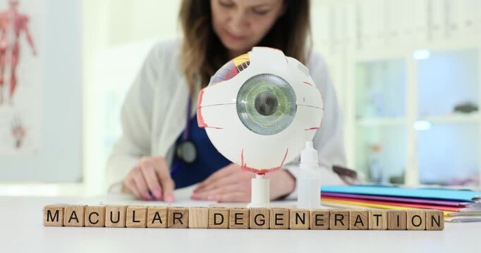 Wooden blocks on desk spell words Macular Degeneration near eye model. Woman ophthalmologist writes chart planning therapy to save central vision