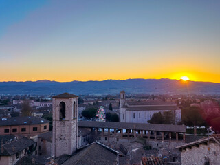 A stunning panoramic view of the medieval town of Gubbio at sunset, with its historic buildings, bell towers, and a distant Ferris wheel, framed by rolling hills