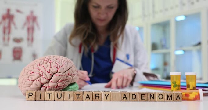 Wooden blocks spell words Pituitary Adenoma by brain model. Woman doctor reads scan results while explaining hormone tumor risk for patient in clinic
