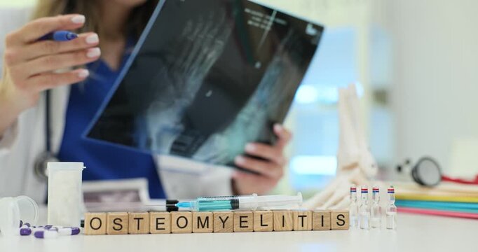 Wooden blocks spell word Osteomyelitis near X-ray image on medical table. Woman doctor holds radiograph film pointing pen toward infected bone area